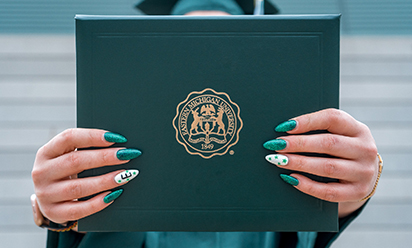 woman holding diploma cover; fingernail polish is green and white and one nail has a large E