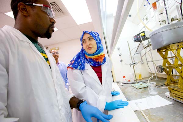 A male and female student in white lab coats work together in a science lab. 