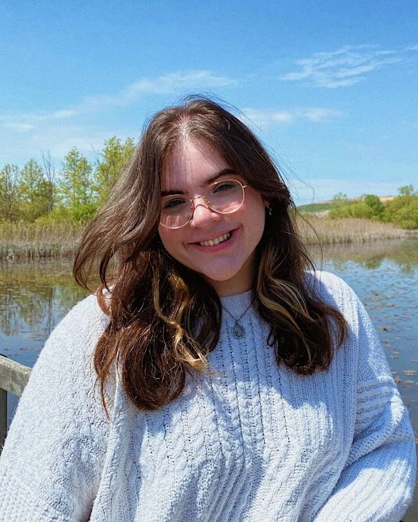 A smiling person with long brown hair wearing glasses and a white sweater stands on a wooden bridge over a body of water with trees in the background.