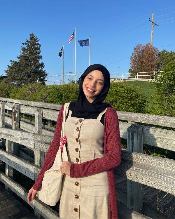 A smiling woman wearing a hijab, khaki jumper, and long-sleeved, maroon shirt stands on a wooden bridge.