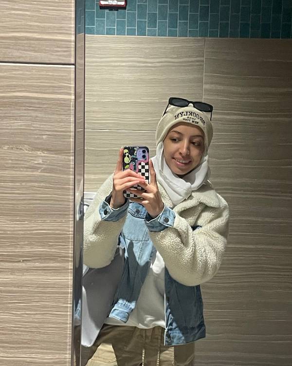 A smiling woman wearing a hijab a cream and denim jacket and khaki-colored pants with sunglesses on her head taking a selfie in a mirror.