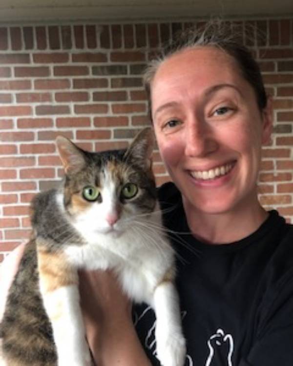 A smiling person with dark hair pulled back from the face wears a dark tee-shirt and holds a white and brown tabby cat.