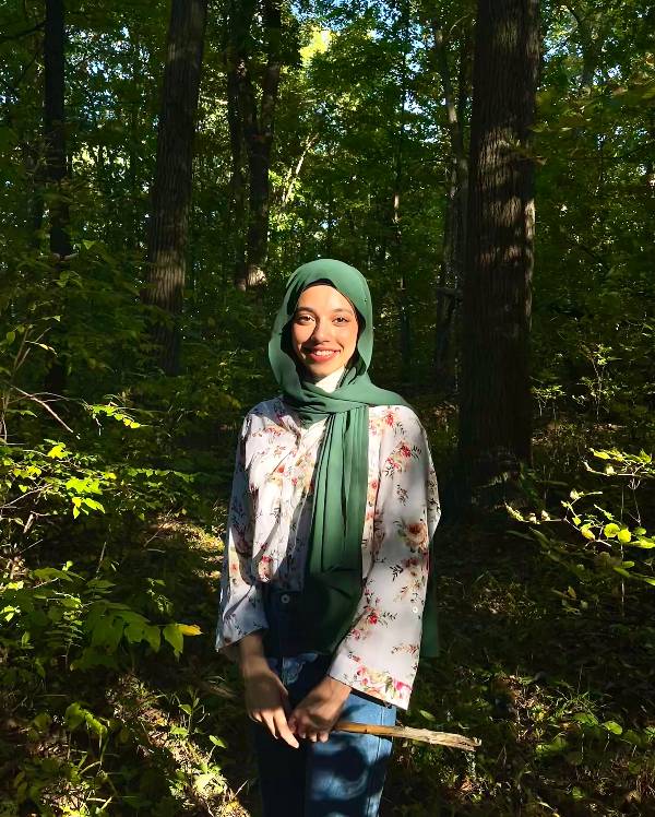 A smiling person wearing a green hijab, flowered blouse, and blue jeans stands in a forest surrounded by greenery.