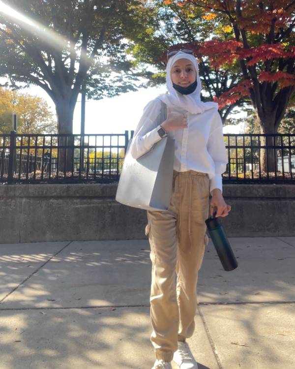 A person wearing a hijab, khaki pants, and a button-down shirt stands outside on an autumn day.