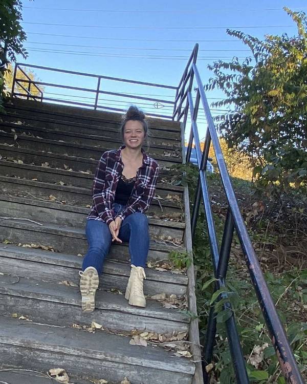A smiling person with brown hair pulled back on their head wears a plaid shirt open over a tank top, jeans, and cream-colored combat boots and sits on an outdoor staircase on a clear day.