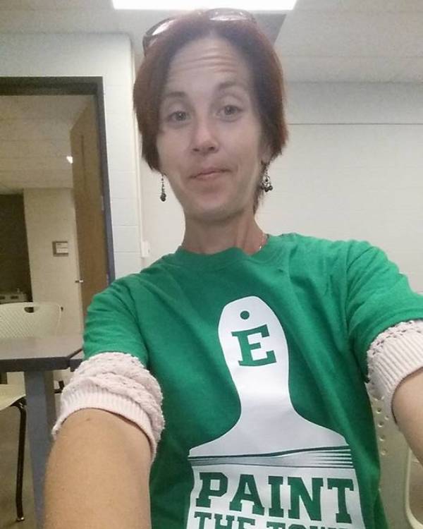 A selfie of a light-skinned person with short red hair wearing an Eastern Michigan tee-shirt and standing in the hallway outside a classroom at EMU.