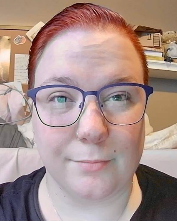 A closeup photo of a smiling person with slicked-back, red hair wearing glasses and a dark top.