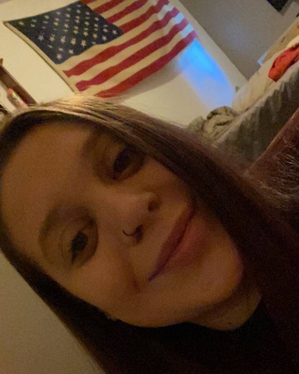A selfie of a smiling person with long, straight brown hair and a nose ring with an American flag hanging on a wall in the background.