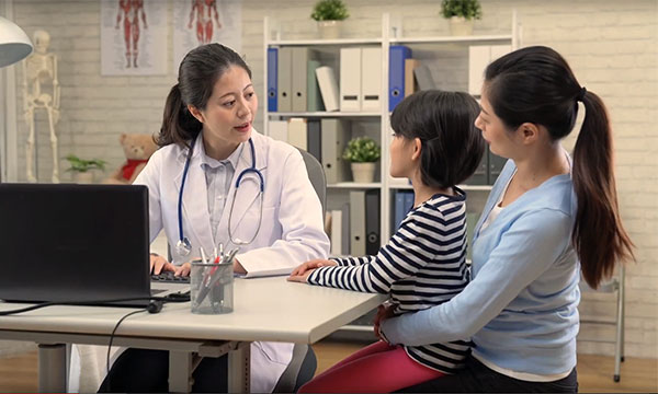 A smiling doctor sitting at a computer talking to a mother and a child.