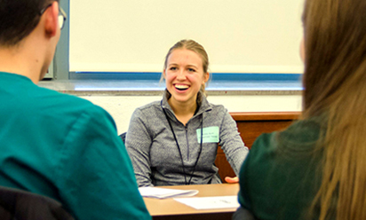 A female student smiles across the table at male and female students.