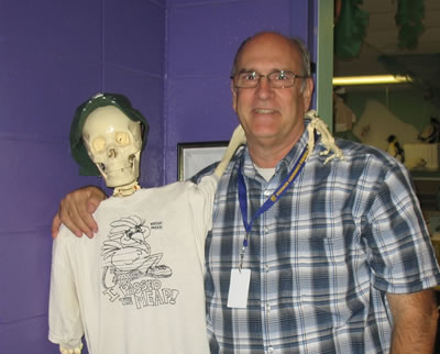 A photo of Gary Koppelman posing with a life-size model of a human skeleton which is earing a hat and a T-shirt that reads, "I passed the MEAP!"