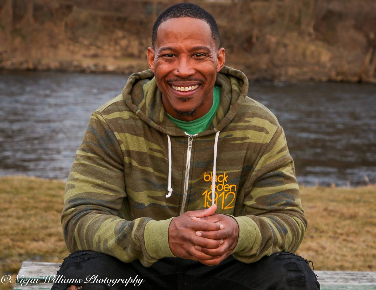 Cozine sits crosslegged with his hands together in front of him. He has a big smile on and is wearing a camoflague hoodie and black pants. He sits outside on some grass and there is a river behind him.