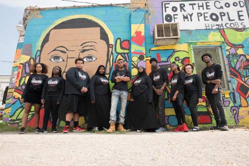 Staying Power Ensemble Posed In Front of a Painted Mural on the side of a brick wall 