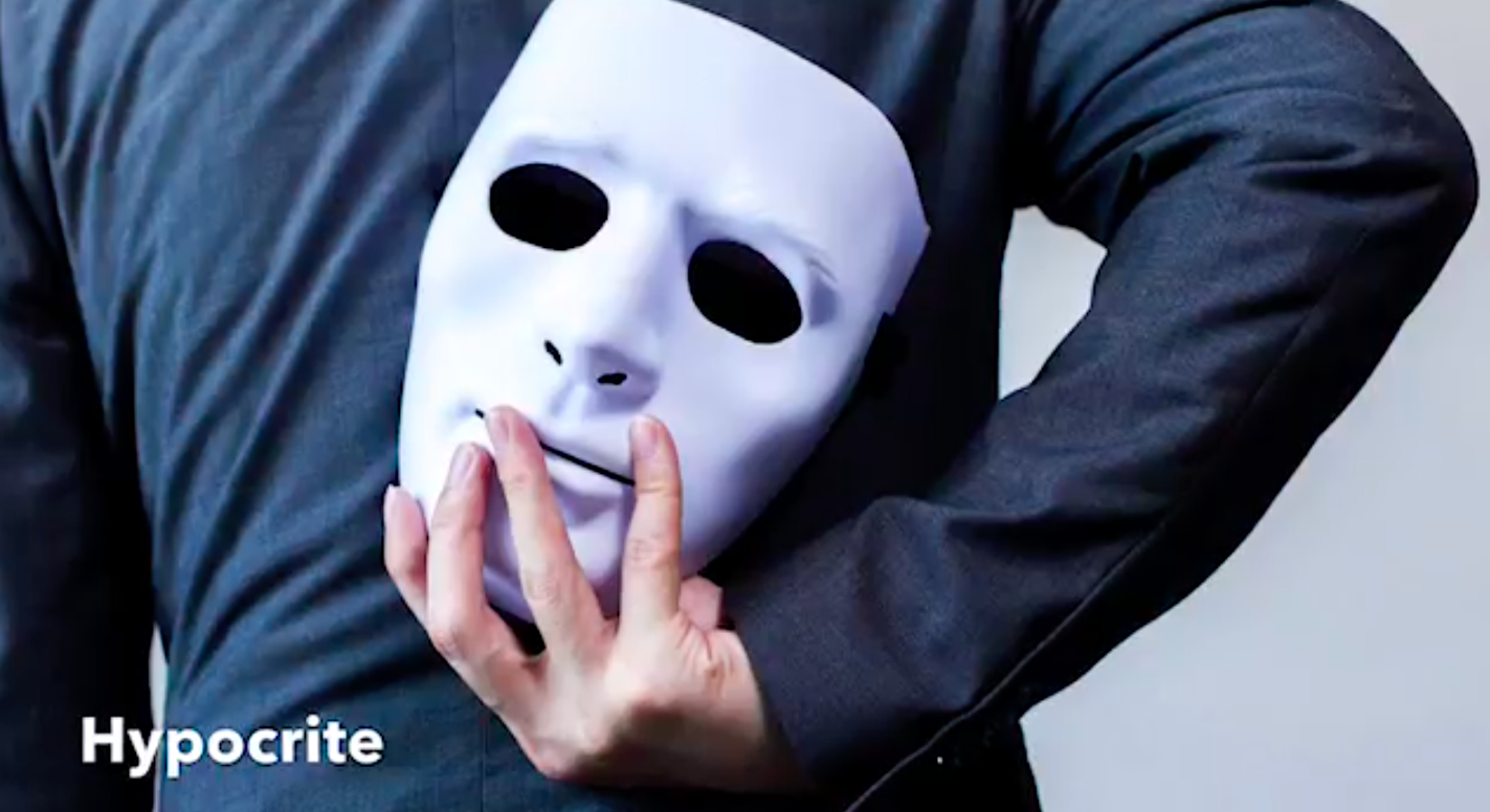 A white person whose face is hidden is wearing a long-sleeved black Tshirt holds a white mask behind their back.