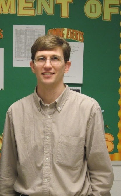 A white man with glasses standing in front of a bulletin board wearing dress clothes.