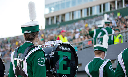 the back of a band member, with a bass drum with block e attached to him; crowd in the background.