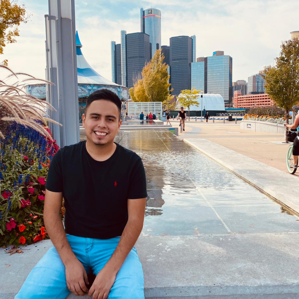 A picture of David sitting on a cement bench at the Detroit Reiverwalk. Behind him is the skyline of Detroit. 